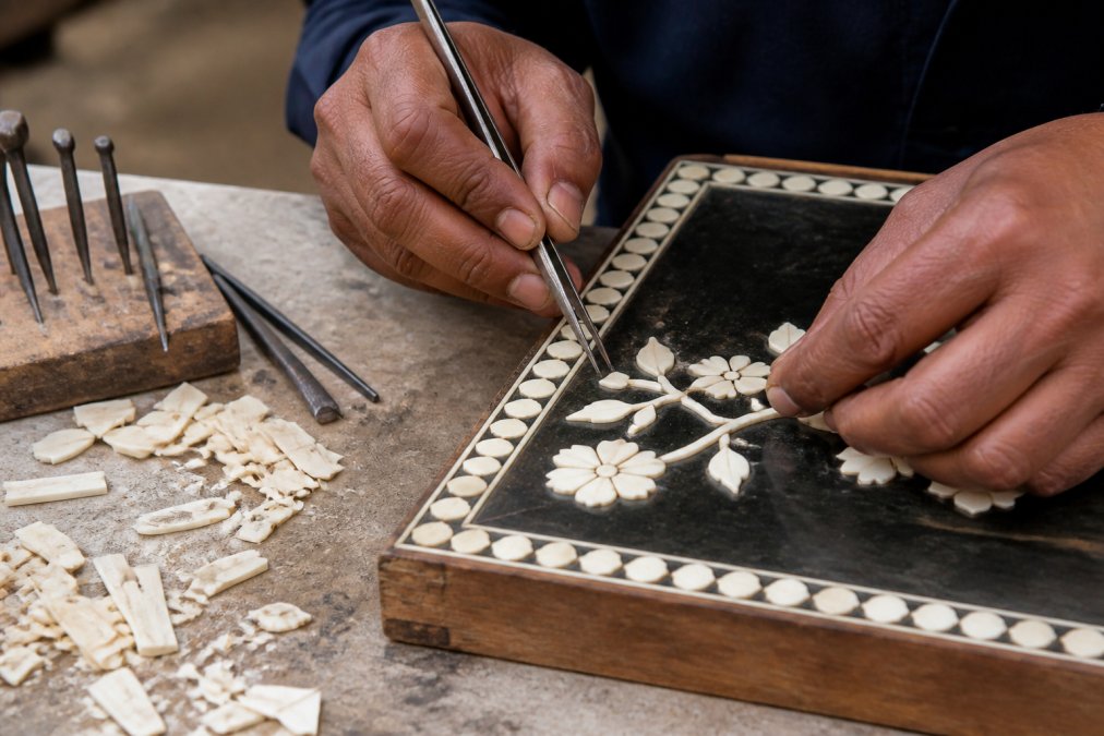 Close-up of an artisan's hands setting small carved bone pieces into a dark resin bed on a timber frame, fine tools and loose bone fragments on the workbench beside them
