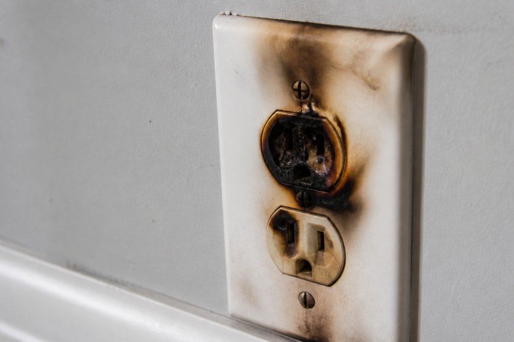 Close-up of a scorched electrical outlet with visible burn marks, blackened discoloration around the plug holes, and charred insulation showing clear signs of electrical damage and arcing