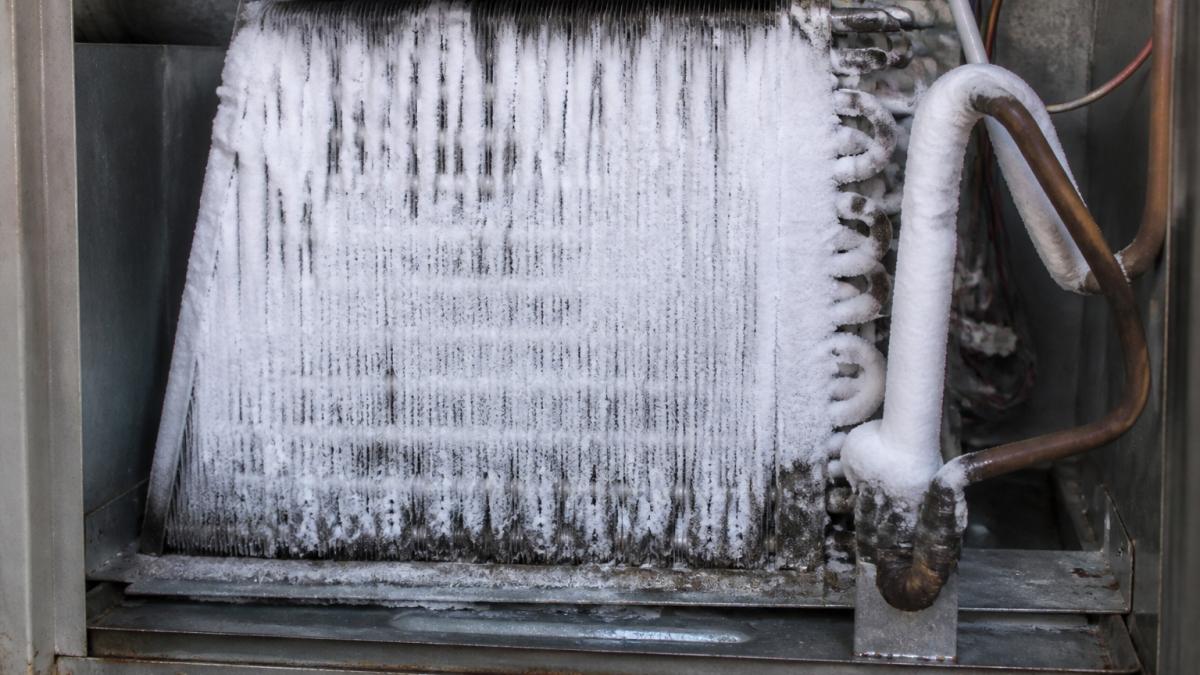 Close-up of a residential AC evaporator coil covered in thick white frost and ice, with refrigerant lines visibly frozen, inside an open air handler cabinet