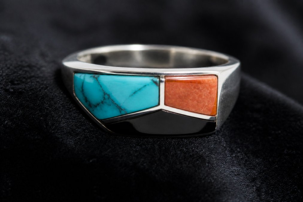 Close-up macro photograph of a Zuni inlay ring resting on dark velvet, showing the smooth flush surface where turquoise, coral, and jet meet the sterling silver channel under soft directional lighting