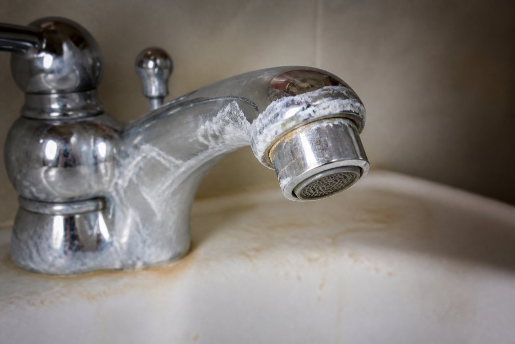 Close-up of bathroom sink faucet with white and chalky limescale deposits coating the chrome aerator and surrounding ceramic surfaces, with hard water staining visible on the sink basin
