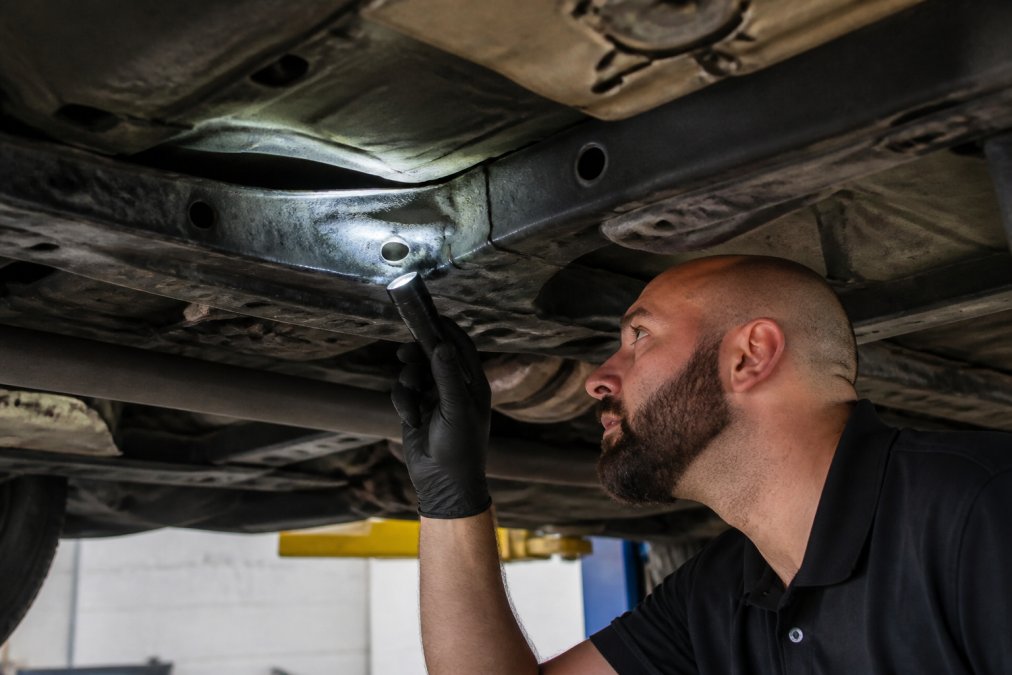 Signs of Hidden Frame Damage: How to Inspect Professional inspector examining vehicle frame rail with flashlight, revealing visible buckling and metal creasing along the undercarriage