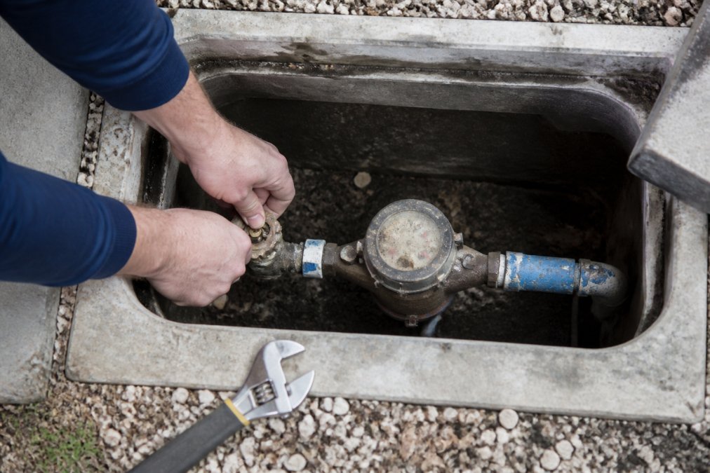 Close-up of a homeowner's hands turning the shut-off valve next to a residential water meter in a utility box, with a wrench resting on the concrete