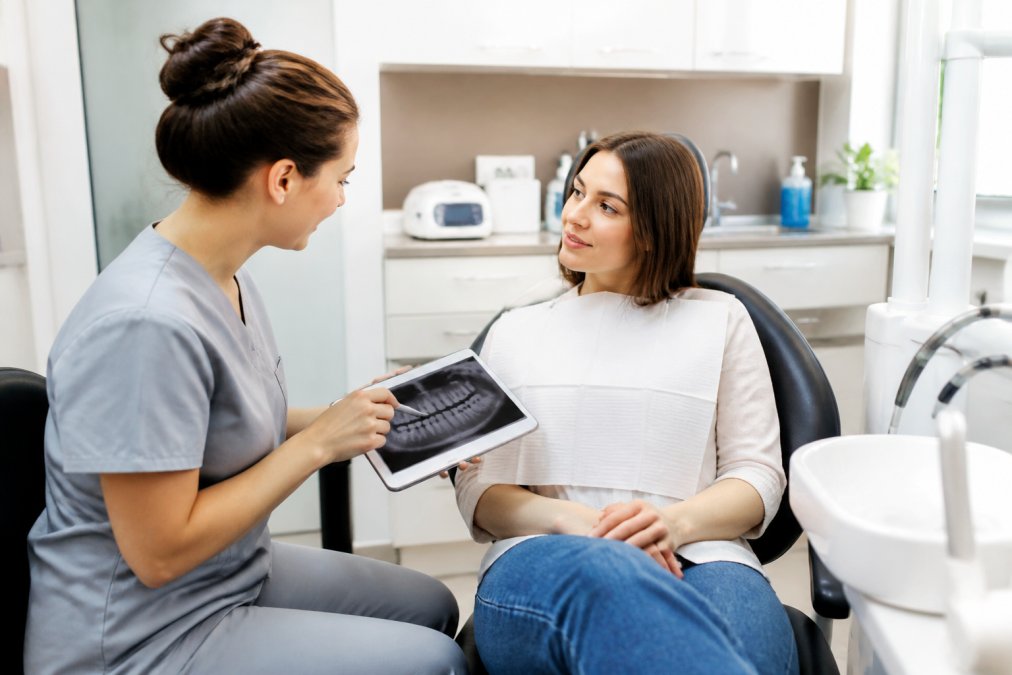 A dental professional in scrubs sitting beside a patient and pointing to a treatment plan displayed on a tablet screen, both engaged in calm conversation in a bright, modern dental office with clean white cabinetry visible in the background