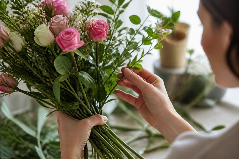 Close-up of hands gently holding a bouquet of fresh flowers, inspecting a green stem and vibrant petals in a bright room