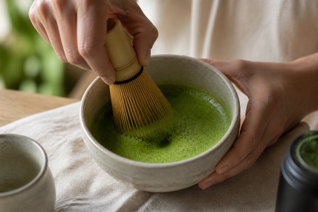 Hands whisking vibrant high quality matcha into a thick, frothy foam inside a ceramic bowl with a bamboo whisk