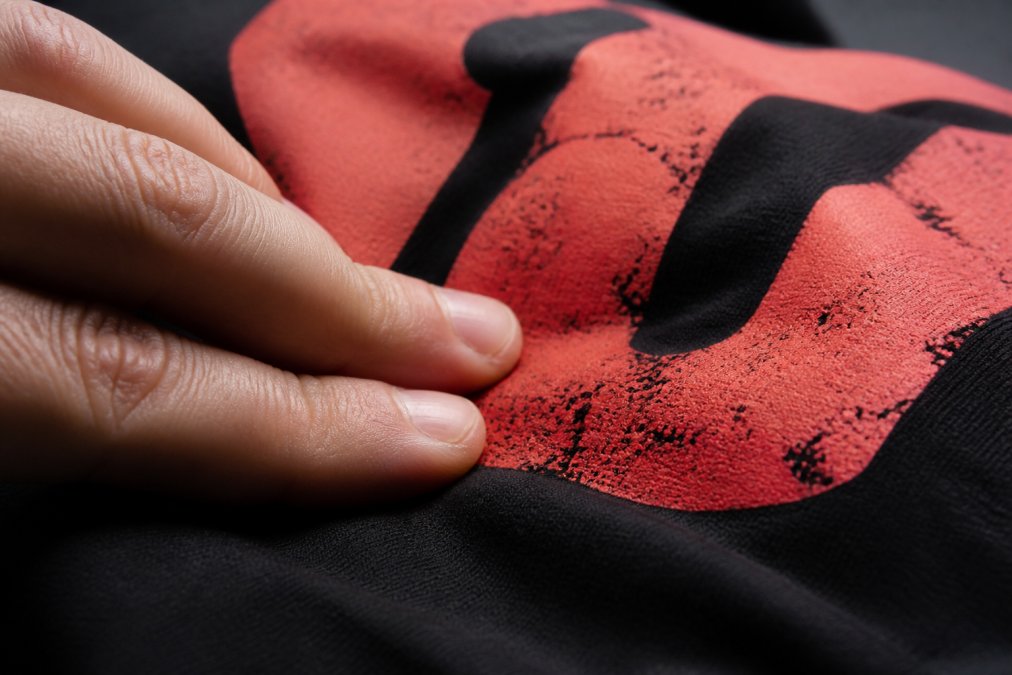 Close-up macro photograph of a person's fingertips pressing into a printed graphic t-shirt, showing the raised ink texture and soft-hand feel of the print surface