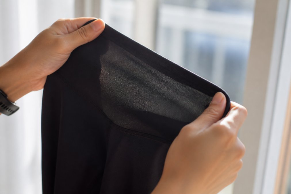 Close-up of hands stretching black athletic leggings against bright window light, testing opacity and elasticity of the fabric
