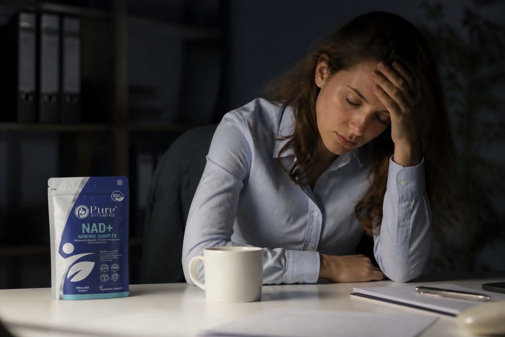 Person at a desk looking fatigued, resting their head on their hand with an empty coffee cup nearby, showing signs of exhaustion and mental fatigue in dim office lighting