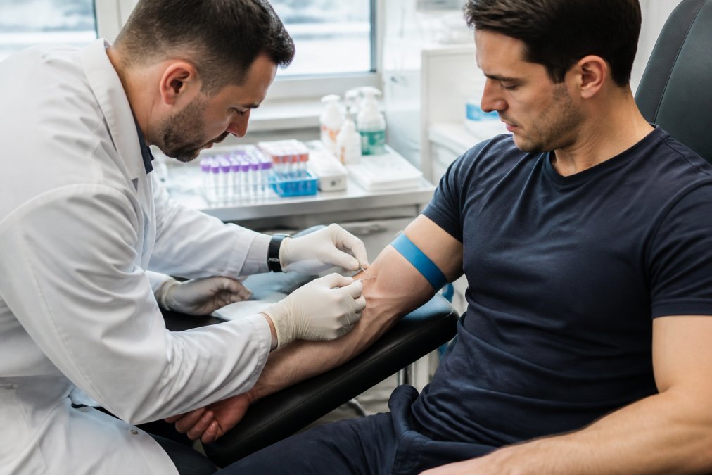 Phlebotomist in white lab coat drawing blood from patient's arm during morning lab visit, patient seated in comfortable chair, bright clinical lighting, focused on needle insertion technique