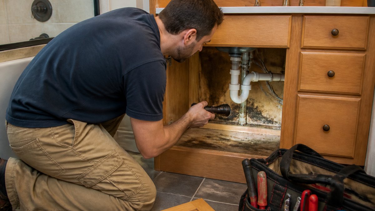 Homeowner using a flashlight to inspect for mold behind bathroom vanity under the sink cabinet pipes