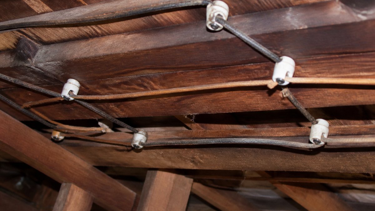 Close-up photograph of aged knob-and-tube wiring running through exposed wooden ceiling joists in an attic, showing white ceramic knobs and cloth-insulated wires