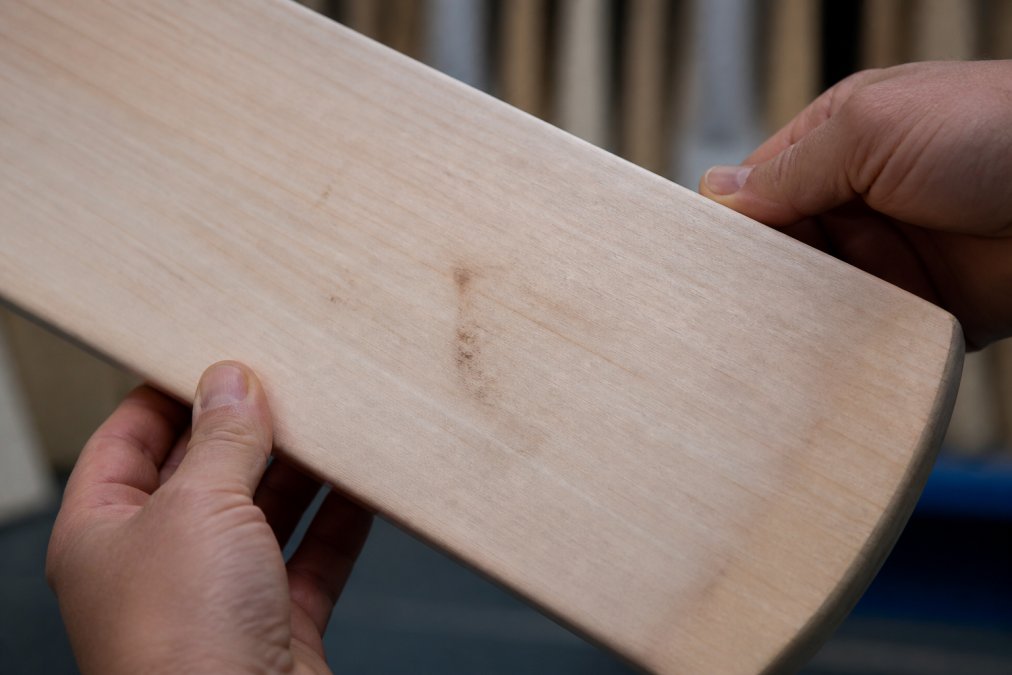 Close-up photograph of a cricket bat face held in two hands under bright light, showing visible willow grain lines and a dark stain mark on the blade