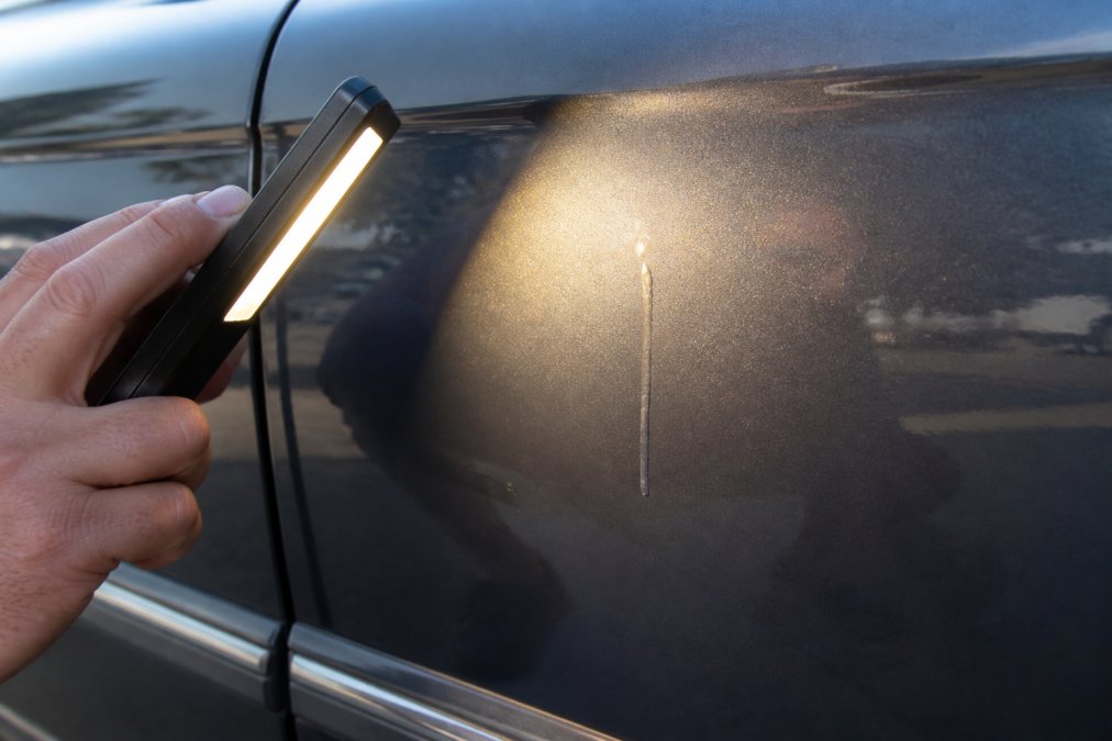 Close-up photograph of a car door panel showing a visible paint drip running down the surface, with a hand holding a bright inspection light at an angle to reveal the flaw