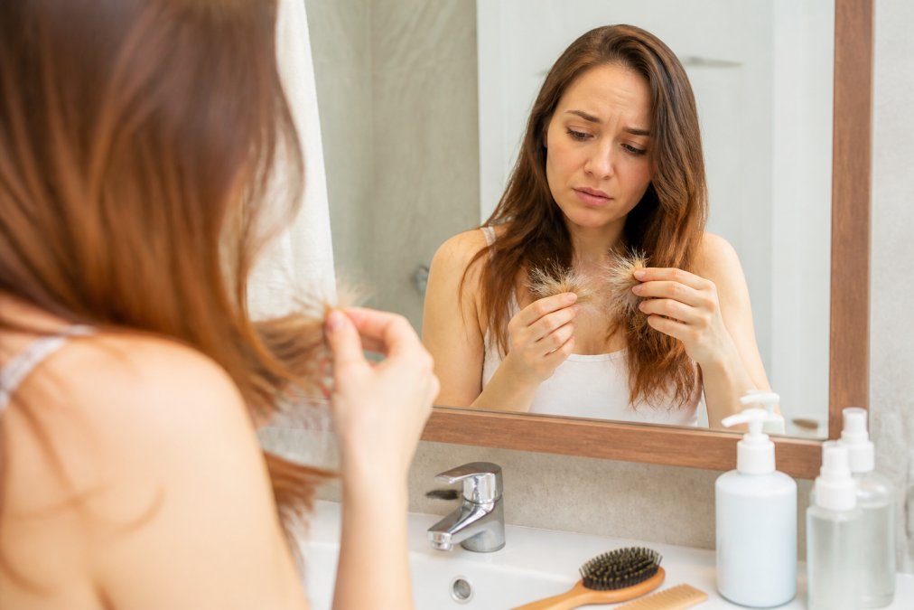Close-up of a woman examining her dry, brittle hair ends in a bathroom mirror, with a comb and hair products on the counter