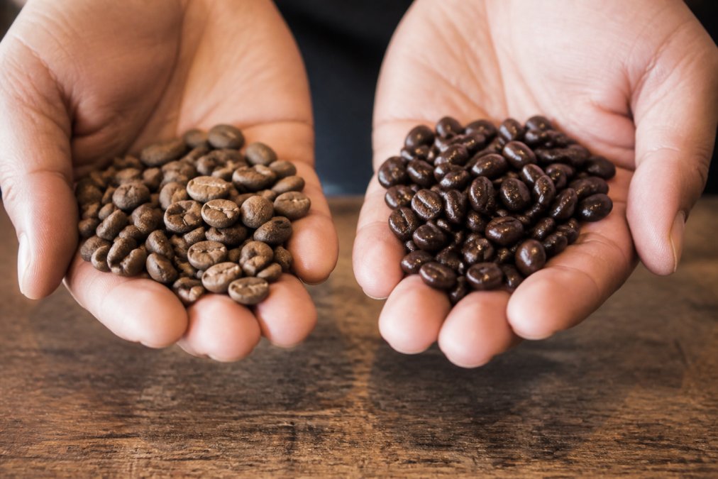 Close-up of two handfuls of coffee beans side by side on a worn wooden surface, one batch visibly dull, dusty, and dry with matte surfaces, the other batch glossy and dark with visible surface oils catching warm kitchen light