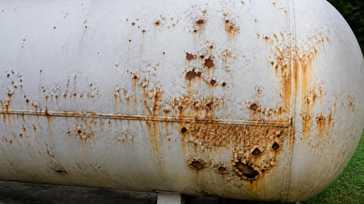 Close-up of propane tank exterior showing rust spots, orange corrosion staining, and visible pitting on the metal surface in natural daylight