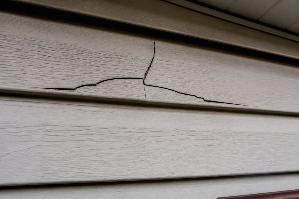 Close-up of house siding showing visible cracks and fractures running through vinyl or fiber cement panels, with natural daylight highlighting the damage and weathered edges
