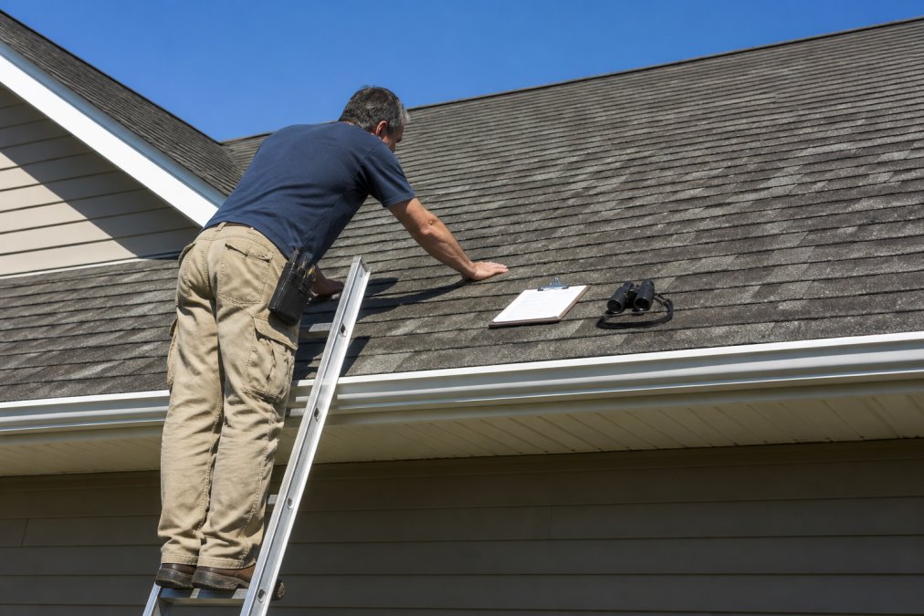 A homeowner standing on a ladder inspecting asphalt shingles on a residential roof, with a clipboard and binoculars nearby, clear blue sky background