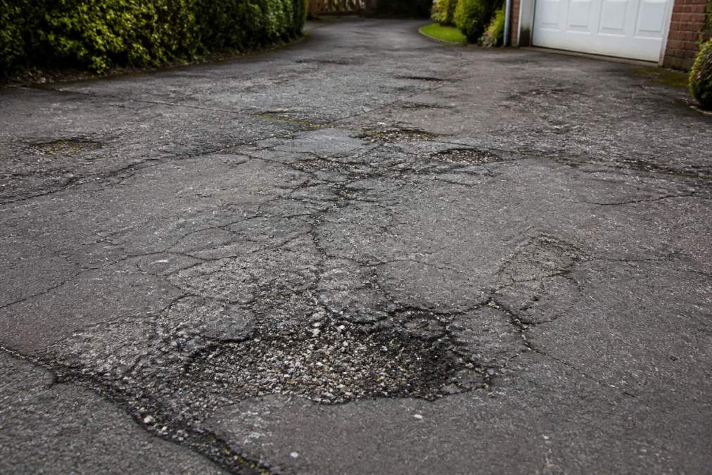 Close-up of a residential driveway showing visible cracks, potholes and deteriorated surface with patches of discolouration