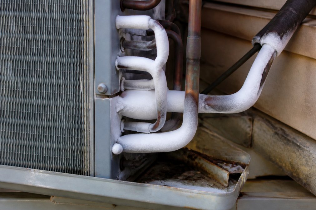 Close-up of outdoor AC condenser unit with visible frost and ice buildup coating the copper refrigerant lines and aluminum fins, showing physical evidence of system pressure loss