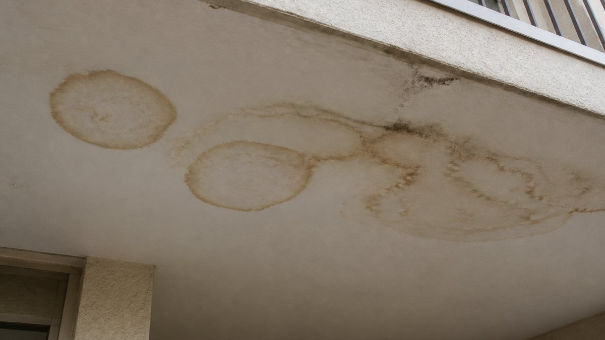 Close-up of water stains and discoloration on ceiling below a balcony, showing brown rings and moisture marks on white drywall with soft lighting