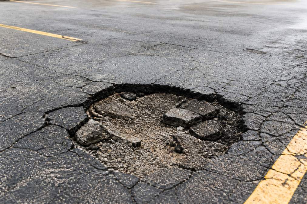 Wide view of a deep pothole in a parking lot showing damaged asphalt edges, exposed base material, and surrounding deteriorated pavement