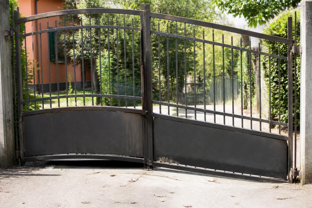 Close-up of a sagging metal driveway gate with visible bowing in the frame and misalignment at the hinges, photographed in daylight
