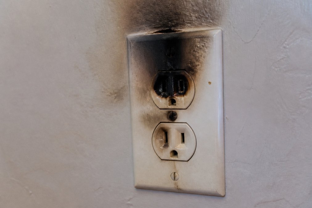 Close-up of a charred electrical outlet with visible scorch marks and discolored wall area surrounding the outlet
