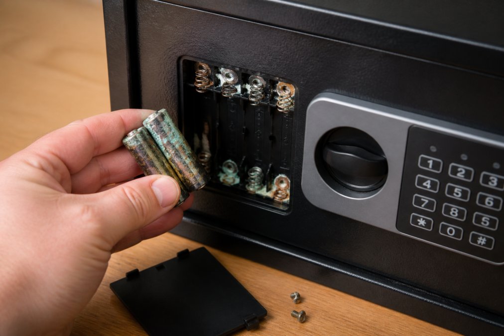 Close-up of a hand carefully removing and inspecting batteries from a digital safe's battery compartment, showing corrosion on the battery terminals and metal contacts