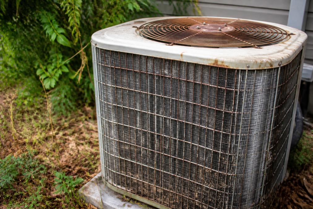 Close-up of an aging HVAC outdoor unit showing rust, dust buildup, and weathered metal fins in a residential backyard with overgrown vegetation nearby