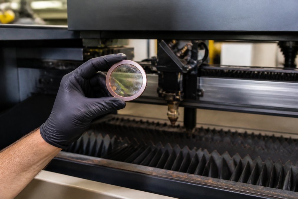 Close-up of a production manager's gloved hand inspecting a laser cutter lens for debris and burn marks under bright workshop lighting