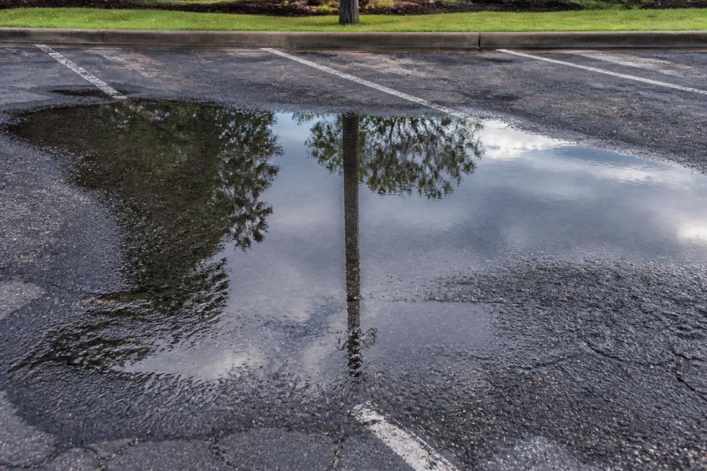 Parking lot surface with visible standing water pooling in low spots, showing poor drainage and water accumulation after rain, with reflections visible in the still water