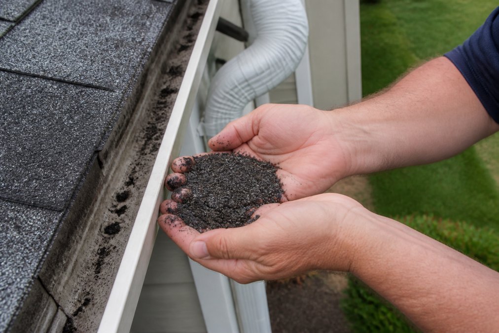 Close-up of a homeowner's hands scooping dark asphalt shingle granules from a residential gutter, with a downspout and roofline visible in the background