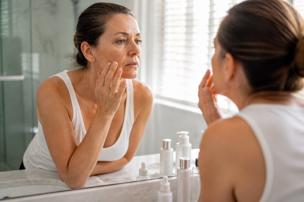 Woman examining her skin in a mirror, one of the signs your skincare routine is wrong, while standing in a bathroom.