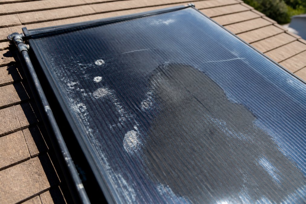 Close-up of a solar pool panel mounted on a residential roof showing visible cracks, white delamination spots, and water staining on the dark surface under bright sunlight
