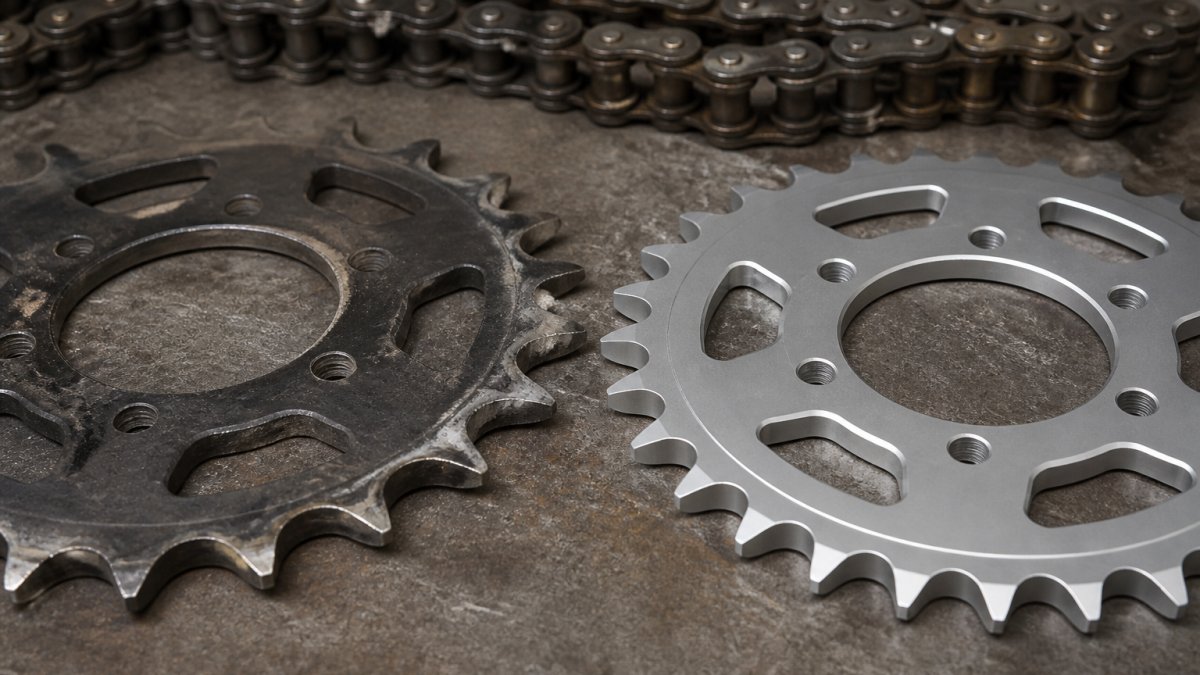 Worn, hooked supermoto sprockets compared to a new sprocket with even teeth on a workshop bench next to a drive chain.