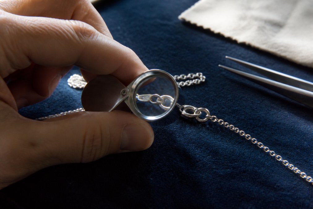 Nahaufnahme einer Hand, die eine feine Silberkette mit einer Juwelierlupe untersucht, um die Punze zu erkennen. Die Kette liegt auf einem dunkelblauen Samttuch, weiches Tageslicht fällt von der Seite ein