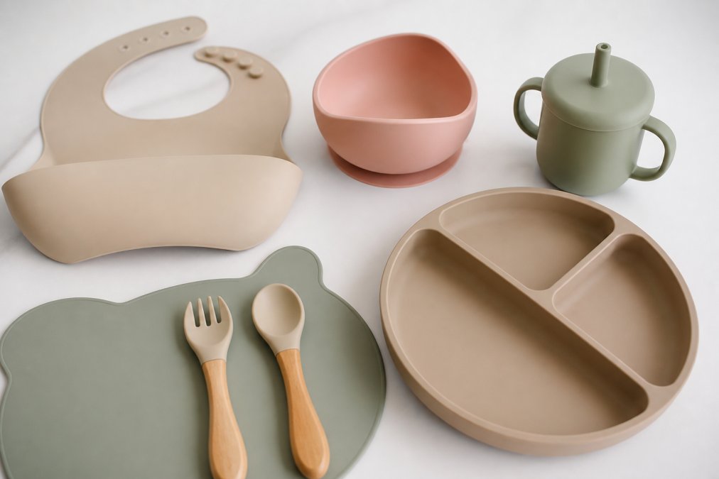 Flat lay of a complete silicone feeding bundle spread on a clean white surface, showing a coordinated bib, suction bowl, suction plate, spoon, fork, cup and placemat in soft pastel colours, natural overhead lighting