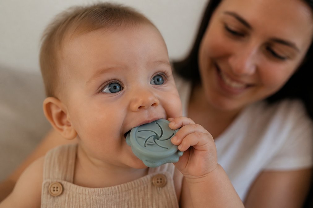Close-up of a teething baby with a soft silicone teether in their mouth, showing visible comfort and relief, with a blurred parent's face smiling in the background and soft natural afternoon lighting