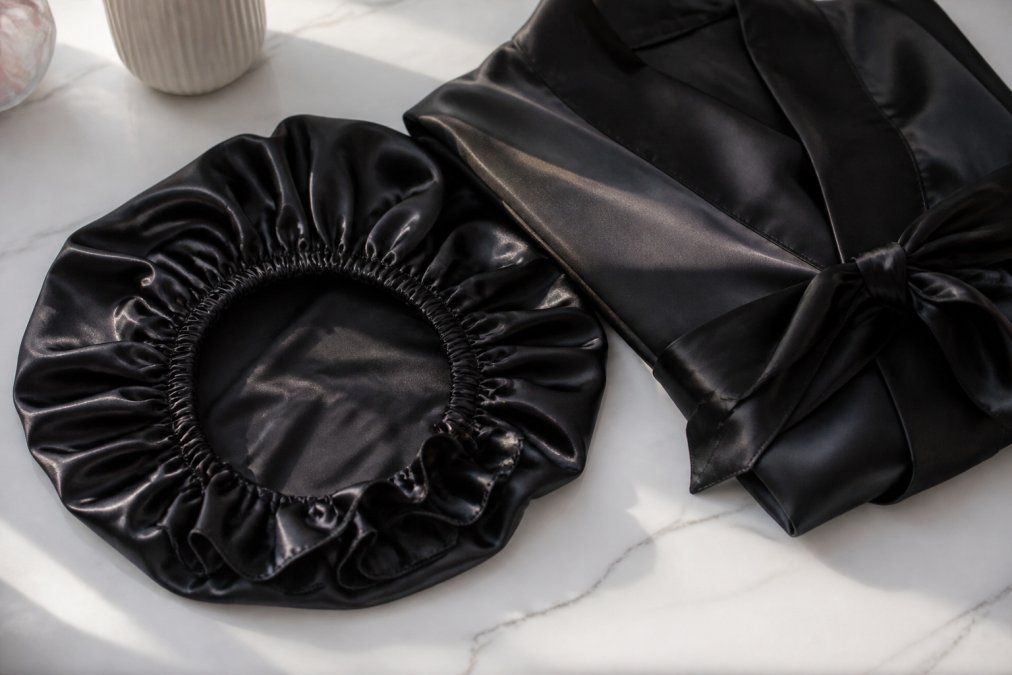 Close-up of The Whispered Crown luxury mulberry silk bonnet in deep Onyx Reign colorway laid flat on a white marble surface alongside a folded silk robe, minimal elegant styling with soft natural light