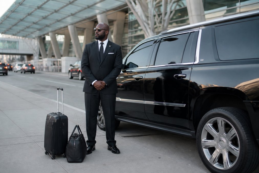 A professional chauffeur in a black suit standing beside a black luxury SUV at a modern airport terminal curb, with luggage nearby and soft daylight