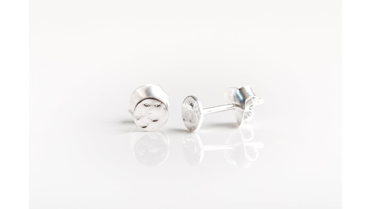 Close-up of a woman's earlobe wearing a tiny sterling silver hammered disc stud earring, soft natural light, showing the delicate texture of the metal against skin