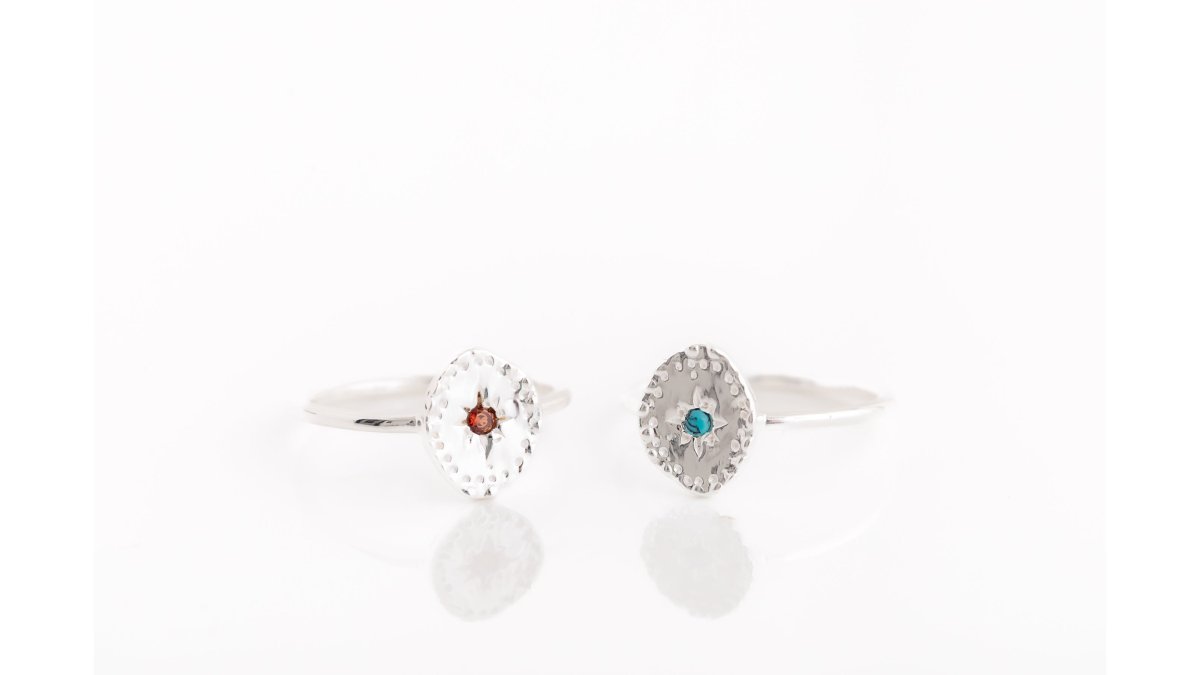 Close-up of multiple handcrafted sterling silver rings displayed on neutral stone surface, showing varied textures, gemstone settings, and hammered finishes catching natural daylight