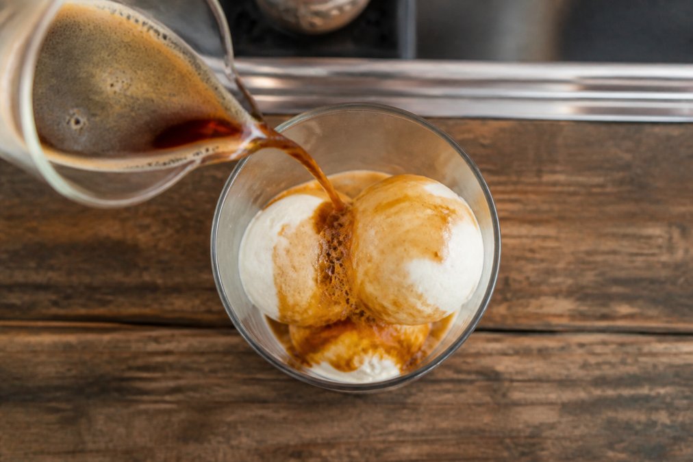 Overhead shot of a chilled glass containing three scoops of vanilla gelato, with a thin stream of hot espresso being poured over the ice cream, showing the rich brown crema mixing with the melting gelato and creating a creamy texture