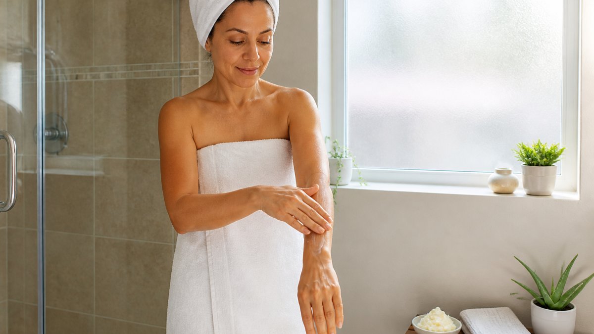 Woman in a towel applying body butter to her arm in a sunlit bathroom