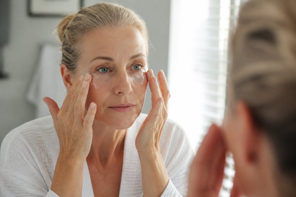 Close-up of a woman in her fifties gently patting a cooling eye gel under her eyes at a bathroom mirror in soft morning light