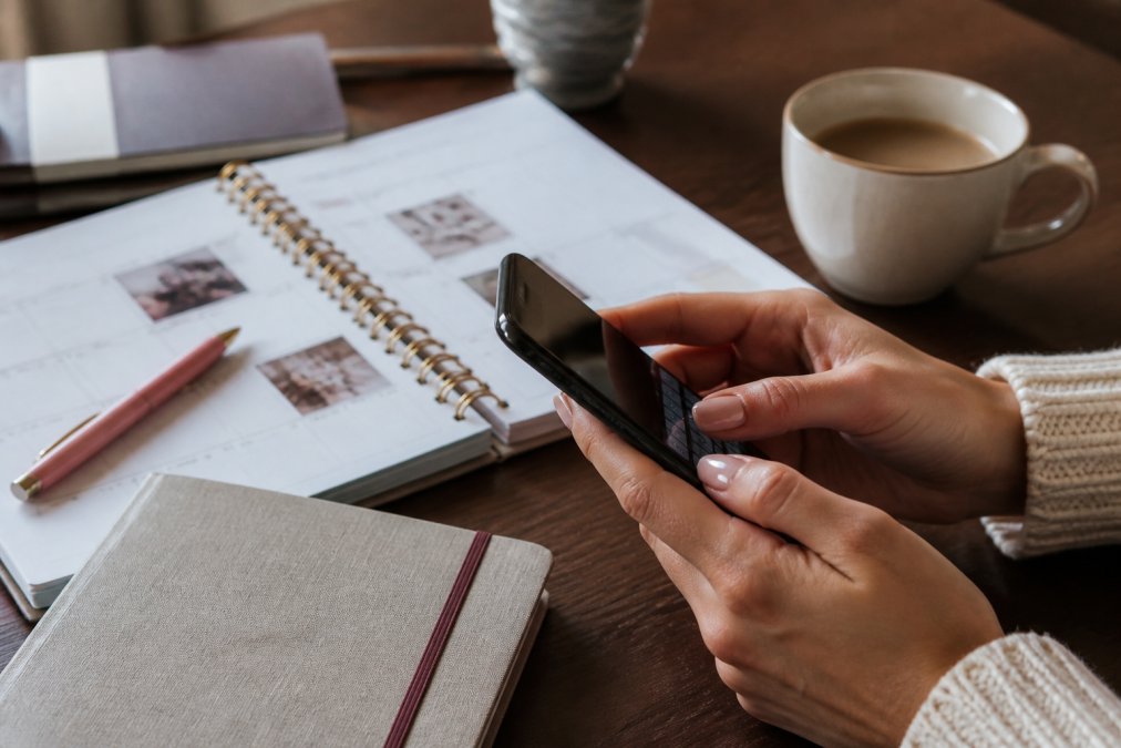 Close-up of hands typing on smartphone keyboard, setting up content calendar and scheduling posts, coffee cup and planner on desk in background
