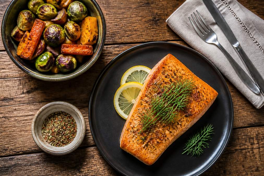 Overhead shot of a rustic wooden table set with a pan-seared salmon fillet topped with fresh dill, alongside a bowl of roasted seasonal vegetables and a small ceramic dish of seasoning blend