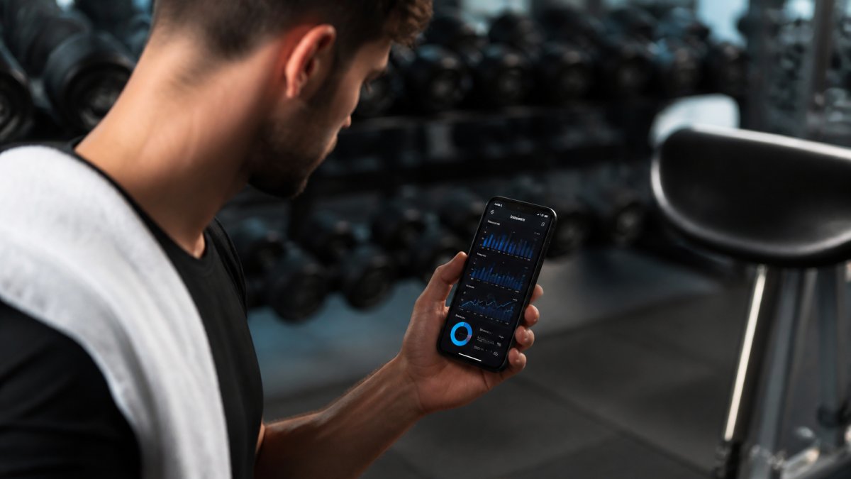 Person at gym holding smartphone displaying workout data with visible progress charts and performance metrics on screen, focused expression reviewing performance